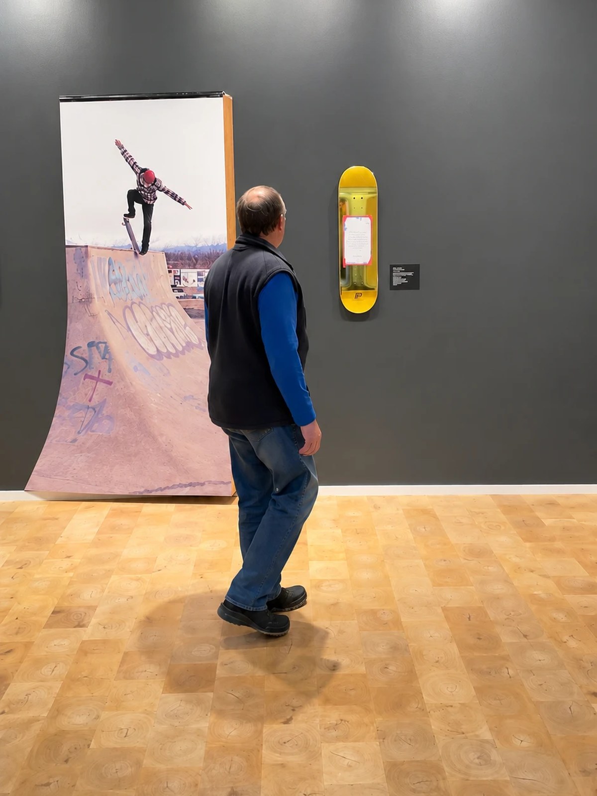 The deck hung in the gallery, a visitor looking along the wall at the skate works beside it