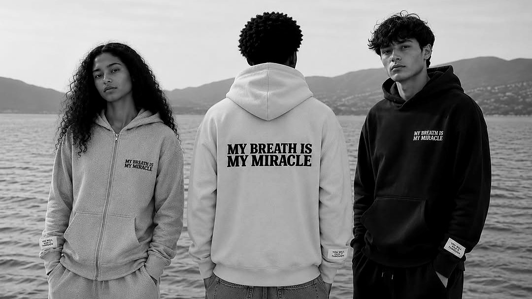 Three people in My Breath Is My Miracle hoodies on a pebble shore, the one in the middle turned to show the print across the back